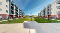 Located at 1913 E 44th St in Joplin, Missouri, this residential community features an expansive outdoor amenity campus designed to offer resort-style living. The grounds are framed by modern, multi-story buildings clad in a distinctive combination of teal, navy, beige, and red brick exteriors, each equipped with private balconies that overlook the shared outdoor spaces below.

At the heart of the community is a generously sized, freeform swimming pool with an adjacent hot tub, surrounded by a wide concrete pool deck furnished with lounge chairs and a covered cabana area supported by natural wood columns. The shaded cabana provides a comfortable retreat from the sun while maintaining views of the pool and surrounding greenery.

Beyond the pool area, well-maintained lawns and concrete walkways connect various amenity zones throughout the complex. A fenced sport court offers an additional recreational outlet, while a dedicated dog park with a gated chain-link enclosure provides a safe space for pets. Seating areas with stylish outdoor furniture are thoughtfully placed along pathways, creating inviting spots for residents to gather and relax.

Outdoor patio spaces adjacent to the buildings feature dining sets with umbrellas, ideal for casual outdoor meals. The overall campus is open, bright, and well-maintained, offering a welcoming community atmosphere under the Missouri sky.