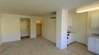 This single-floor studio is designed with an efficient, open-concept layout that maximizes the use of available space. The main living area is finished with neutral beige carpeting that extends throughout, creating a cohesive and understated aesthetic. A wall-mounted mini-split air conditioning unit is positioned near the ceiling, providing climate control without occupying floor space.

A structural partial-wall divides the living area from the kitchen alcove, which features white cabinetry, a white gas range, a compact sink, and tile backsplash. The white finishes throughout the kitchen contribute to a clean, bright appearance.

Adjacent to the entry door, a built-in open closet offers dedicated storage space, making practical use of the area near the entrance. The front door is solid with standard hardware, providing a straightforward entry point to the unit.

The overall color palette is neutral — white walls, light-toned carpet, and white fixtures — lending the space a simple, functional character. The single-floor layout ensures ease of movement throughout, making the studio well-suited for straightforward, everyday living.