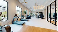 Located at 3558 Elm St in Atlanta, GA, this well-appointed fitness center offers a generously sized, single-floor exercise facility designed to support a full range of workout routines. The space is thoughtfully organized into distinct zones, each optimized for a specific training focus.

The cardio area lines the perimeter with large floor-to-ceiling windows that flood the space with natural light and provide views of the surrounding greenery outdoors. Multiple treadmills and elliptical machines are arranged along this naturally lit wall, complemented by potted fiddle-leaf fig trees that add a refreshing, organic touch to the environment.

The strength training zone is centrally positioned and features a professional-grade power rack with an integrated weight and accessory storage system, a full dumbbell rack, cable machines, and various benches. Large mirrors enhance the sense of openness while supporting proper form during workouts.

A standout feature is the glass-enclosed cycling studio, separated from the main floor by sleek black-framed glass panels. Outfitted with stationary bikes and textured accent walls, it provides a dedicated, boutique-style space for spin sessions.

Overhead, coffered ceilings with warm recessed lighting panels and multiple ceiling fans create a bright yet comfortable ambiance. The light-toned flooring and neutral wall palette maintain a clean, modern aesthetic throughout, making this fitness center both functional and visually inviting.