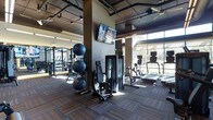 This fitness center offers a comprehensive workout environment designed to accommodate a wide range of exercise needs. The space features an industrial-inspired aesthetic, with exposed ceiling infrastructure, structural columns, and ceiling fans that contribute to an open, loft-like atmosphere. Dark, patterned carpet tiles cover the floor throughout, lending a modern and practical finish to the workout area.

The facility is well-stocked with a diverse selection of equipment. Strength training options include cable and resistance machines, a multi-station weight rack system, and a set of stability balls stored on a dedicated rack. The cardio section is positioned along the perimeter near expansive floor-to-ceiling windows, which flood the space with natural light and offer views of the surrounding outdoor landscape. Equipment here includes treadmills, rowing machines, and upright cardio units.

Wall-mounted flat-screen televisions are positioned throughout for entertainment during workouts. The generous ceiling height enhances the sense of openness and airflow, while the large glazed window wall creates a bright, energizing environment. The overall layout is thoughtfully organized, allowing for comfortable movement between stations and a functional, distraction-free training experience.