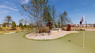 Located at Liberty Grand Boulevard in Powell, Ohio, this outdoor amenity space is centered around a thoughtfully designed putting green that serves as a standout recreational feature of the community. The putting green spans a generous area with smooth, high-quality synthetic turf, multiple pin placements, and strategically placed sand bunkers that replicate an authentic golf course experience. A central island landscape feature adds visual interest and challenge to the green, showcasing a combination of mature trees, ornamental grasses, decorative river rock, and large natural boulders. The surrounding grounds are meticulously landscaped with manicured turf, evergreen shrubs, flowering plants, and young deciduous trees that provide seasonal color and texture. Winding concrete pathways connect the putting green to adjacent amenities, including tennis and pickleball courts visible in the background. A community clubhouse or pool facility with an iron perimeter fence can be seen further along the path, complementing the overall amenity offering. Elegant lampost-style lighting is positioned throughout the space, extending usability into the evening hours. The entire area enjoys open skies and expansive views, creating a bright, welcoming outdoor environment ideal for recreation and leisure.