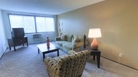 This single-floor space is designed with an open-concept layout that connects the living area seamlessly to the dining space, creating a functional and cohesive flow. The living room is carpeted and features warm, earthy-toned walls complemented by a textured ceiling. A large window with vertical blinds allows natural light to fill the room, enhancing its comfortable atmosphere. The space accommodates a plush sofa and patterned accent chair arranged around a dark wood coffee table, creating a welcoming seating area.

Transitioning from the living room, a distinctly painted accent wall in a rich orange hue marks the entrance to the dining area. This space features a ceiling fan with a light fixture overhead and is furnished with a dining table and chairs, making it suitable for everyday meals. Decorative wall art adds a personalized touch to the dining space.

Throughout the unit, neutral carpet flooring unifies the rooms, while the combination of warm and neutral tones on the walls contributes to a cozy, lived-in ambiance. The layout is practical and well-proportioned, offering clearly defined areas for living and dining within a single connected floor plan.