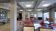 This single-floor space presents a well-designed communal area that balances functionality with contemporary aesthetics. The ceiling is a standout architectural feature, showcasing a coffered design with flush-mounted rectangular light panels and recessed can lighting that together provide even, welcoming illumination throughout the room. Structural columns are integrated cleanly into the layout, contributing to the space's defined zones without interrupting the open flow.

The seating area is furnished with multiple clusters of tables and chairs in a bold coral-red and warm neutral palette, offering flexible arrangements suitable for casual meetings, dining, or social gatherings. Carpet tiles in a subtle pattern cover much of the floor, providing both comfort and visual texture.

Along the perimeter, a reception or service counter is visible, suggesting a staffed amenity or check-in function. A colorful abstract artwork anchors one wall, adding a creative focal point. Glass-paneled doors and interior windows allow views into adjacent spaces, enhancing the sense of openness. Natural light filters in from one side, complementing the artificial lighting. Overall, the space exudes a clean, organized, and inviting atmosphere suited for community use.
