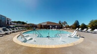 Located in Denver, North Carolina, this single-story community amenity facility at Beckstead Court offers a thoughtfully designed outdoor living experience. The exterior showcases a charming combination of bold blue siding and brick wainscoting, giving the building a modern yet grounded aesthetic.

At the heart of the space is a generously sized resort-style swimming pool, featuring built-in water fountains and in-pool lounge chairs that invite relaxation. The pool deck is lined with additional chaise lounges and is enclosed by a sleek black iron fence, ensuring both safety and visual appeal under the open Carolina sky.

A covered outdoor lounge area extends from the main building, finished with a warm wood-plank ceiling and ceiling fans to keep the space comfortable year-round. This patio is equipped with a built-in linear fireplace set against a brick accent wall and furnished with stylish wicker lounge seating arranged over a patterned area rug, creating an inviting gathering spot. Floor-to-ceiling glass sliding doors connect this covered porch directly to the pool area, maximizing views and airflow.

Entry points include glass-paneled doors framed in sleek dark metal, offering a preview of the amenity space within. A gated iron entryway with brick pillar supports provides a grand, secure passage from the building to the pool deck, completing the seamless indoor-outdoor flow of this community destination.