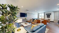 Located at 4121 Concord Creek Place in Richmond, VA, this single-floor community lounge is thoughtfully designed to serve as a welcoming social hub. The space is divided into two functional areas that flow seamlessly together, united by warm hardwood-style flooring and recessed lighting throughout.

The main lounge area is anchored by a striking accent wall featuring a soft, panoramic landscape mural in pastel hues of blue, blush, and gold. A large wall-mounted flat-screen TV takes center stage, flanked by lush indoor plants and tasteful artwork. Comfortable blue leather sofas and tan leather armchairs are arranged around a marble-top coffee table, creating an inviting setting for relaxation or casual gatherings.

The secondary zone transitions into a vibrant entertainment and casual dining area defined by bold cobalt blue accent walls. This section includes a small dining table with chairs, built-in white cabinetry, and cozy seating clusters with gold-toned accent tables. Elevator access is conveniently located here, along with abundant natural light streaming through large windows dressed with striped curtains.

A dedicated game corner features a shuffleboard table, a wall-mounted Connect Four game, and additional lounge seating, adding a playful dimension to the space. The overall ambiance is modern, energetic, and community-focused.