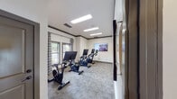 Located at 4095 Lawson Ridge Dr in Huntsville, Alabama, this single-floor spin and cycling studio is a purpose-built fitness space designed to support an engaging and motivating workout experience. The room is outfitted with a row of Echelon stationary bikes, each equipped with individual digital display screens, allowing riders to follow interactive workout programs at their own pace. A large wall-mounted flat-screen television is positioned at the front of the room, providing a shared visual focal point for group cycling sessions or individual entertainment. The studio features neutral-toned carpet flooring with a subtle geometric pattern, contributing to a clean and comfortable atmosphere. Crisp white walls are accented by dark wood crown molding, lending the space a polished, upscale feel. Natural light filters in through several windows dressed with patterned curtains, while recessed panel lighting in the ceiling ensures consistent illumination throughout. Adding a refined artistic touch, large-scale abstract paintings are displayed prominently along the walls, elevating the aesthetic beyond a typical fitness room. A well-lit hallway with light wood-look flooring and additional curated wall art connects this studio to the rest of the facility, reinforcing the thoughtful and cohesive design found throughout the property.