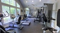 Located at 3004 Dorner Cir in Raleigh, NC, this dedicated fitness center is thoughtfully designed to support a comprehensive workout experience. The space is anchored by soaring vaulted ceilings that lend an open, airy feel, while a ceiling fan promotes comfortable air circulation during exercise. Large, multi-pane windows line the walls, flooding the room with natural light and offering pleasant views of the surrounding greenery, creating an inviting and energizing atmosphere.

The facility is outfitted with a full suite of professional-grade TRUE Fitness equipment, including stationary recumbent and upright bikes, a treadmill, an elliptical trainer, and a multi-station cable strength machine. A dumbbell rack stocked with a range of free weights offers versatility for resistance training. Protective rubber flooring covers the entire workout area, providing safety and durability underfoot.

A full-length wall mirror reflects both the equipment and the natural light, enhancing the sense of spaciousness while allowing users to monitor their form. Recessed lighting and pendant fixtures supplement the natural light for evening workouts. The single-story layout and dedicated entrance make the fitness center easily accessible, offering a focused, private environment for residents to meet their health and wellness goals year-round.