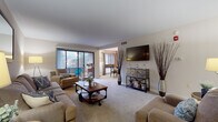 This single-floor residence offers a well-organized layout that balances open communal spaces with comfortable private retreats. The living room serves as the heart of the home, featuring plush carpet, neutral walls, and a wall-mounted flat-screen television above a rustic console. A sliding glass door fills the space with natural light and provides access to an outdoor balcony with wooded views. The living area flows seamlessly into the dining space, which is equipped with a ceiling fan, a round table, and access to the kitchen via a peninsula counter with bar seating and dark granite-style countertops. The kitchen itself features white cabinetry, matching dark granite surfaces, tile flooring, and black appliances, including a gas range and full-size refrigerator. Two carpeted bedrooms offer a calm retreat; both are generously proportioned to accommodate large beds and furniture, with one featuring an open closet with built-in shelving. The bathroom includes a full tub-shower combination with white tile surround. Throughout the residence, a cohesive neutral palette and soft carpeting create a warm, comfortable ambiance.
