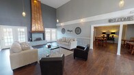 Located at 2095 Valley Greene Dr in Dayton, Ohio, this single-floor community space combines contemporary design with warm, inviting character. The centerpiece of the main lounge is a dramatic copper-clad fireplace that extends toward the soaring double-height ceiling, commanding attention from every angle. Rich dark hardwood flooring runs throughout, complementing the cool grey-toned walls accented by crisp white wainscoting and picture-frame wall molding. Pendant lighting adds a refined touch to the grand vertical space. Large French doors flood the lounge with natural light and provide views to the exterior, while a loft-style upper railing overlooks the main floor, adding architectural depth. The seating area is spacious and well-appointed, flowing seamlessly into a dining area furnished with multiple wooden tables and chairs. A wall-mounted television and a service counter make this area well-suited for gatherings and events. A galley-style kitchen offers ample cabinetry, dark countertops, and full appliances including a refrigerator and dishwasher, supporting a range of catering needs. Throughout the space, decorative wall art and thoughtful lighting create a polished, community-focused ambiance ideal for residents and events alike.