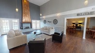Located at 2095 Valley Greene Dr in Dayton, Ohio, this single-floor community space combines contemporary design with warm, inviting character. The centerpiece of the main lounge is a dramatic copper-clad fireplace that extends toward the soaring double-height ceiling, commanding attention from every angle. Rich dark hardwood flooring runs throughout, complementing the cool grey-toned walls accented by crisp white wainscoting and picture-frame wall molding. Pendant lighting adds a refined touch to the grand vertical space. Large French doors flood the lounge with natural light and provide views to the exterior, while a loft-style upper railing overlooks the main floor, adding architectural depth. The seating area is spacious and well-appointed, flowing seamlessly into a dining area furnished with multiple wooden tables and chairs. A wall-mounted television and a service counter make this area well-suited for gatherings and events. A galley-style kitchen offers ample cabinetry, dark countertops, and full appliances including a refrigerator and dishwasher, supporting a range of catering needs. Throughout the space, decorative wall art and thoughtful lighting create a polished, community-focused ambiance ideal for residents and events alike.