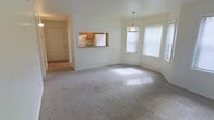 This single-floor space offers a bright, open layout with a neutral color palette and plush carpeted flooring throughout the main living area. A key architectural feature is the pass-through window connecting the living space to the kitchen, providing visual openness while maintaining defined areas. The kitchen, visible through the pass-through, includes wooden cabinetry, a white refrigerator, and a range, offering functional efficiency in a compact footprint. A bay window configuration on one wall floods the living area with natural light, creating a warm and inviting atmosphere, with a pendant light fixture centered nearby adding a subtle decorative touch. The entry area, visible in the background, transitions from the carpeted living space to hard flooring, providing a practical foyer zone. The textured ceiling and clean white walls contribute to a simple, well-maintained aesthetic. The overall layout flows smoothly from entry to living area to kitchen, making the space feel connected and easy to navigate.