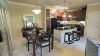 This single-floor residence offers an open-concept layout that seamlessly connects the kitchen and dining areas, creating a cohesive and inviting living environment. The kitchen is a standout feature, showcasing rich dark wood cabinetry that extends to the upper walls, complemented by dark granite countertops and a breakfast bar with sleek bar stools — ideal for casual dining or entertaining. Black appliances, including a refrigerator, microwave, and range, add a modern, unified aesthetic, while pendant lighting and a track light fixture illuminate the space with both function and style. Crown molding along the ceiling adds a refined architectural detail throughout. The dining area flows naturally from the kitchen, anchored by a dark wood dining table with upholstered chairs and a classic pendant light overhead. A large mirror on the dining room wall enhances the sense of space and reflects natural light filtering in from nearby windows. Neutral-toned walls and plush carpeting throughout the living areas create a warm, welcoming ambiance, tying together the space's contemporary design elements with understated elegance.