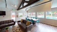 This single-floor space blends industrial and contemporary design elements to create a welcoming and functional community environment. The interior is immediately distinguished by its soaring vaulted ceilings, accented by dark-stained exposed wooden beams and visible ductwork that lend the space an urban, loft-inspired aesthetic. Durable wood-plank flooring runs throughout, tying each area together cohesively.

The central lounge area serves as the heart of the space, anchored by a striking floor-to-ceiling stacked stone fireplace with a sleek linear insert. Comfortable dark leather sofas and accent chairs are arranged around colorful patterned area rugs, creating inviting seating clusters for gathering and relaxation. A wall-mounted television provides an additional entertainment focal point.

Adjacent to the lounge, a dining area features a large communal table surrounded by vibrant teal chairs, positioned beneath a bank of windows that fill the room with natural light and offer views of an outdoor pool and surrounding grounds. Track lighting along the beamed ceiling supplements natural illumination throughout.

Toward the back of the space, a professional leasing or administrative area features glass-paneled office partitions and branded signage, clearly delineating workspace from community areas. The overall ambiance is modern, energetic, and purposefully designed for both social interaction and everyday utility.