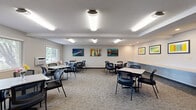 Located at 2221 Hanley Rd in Hudson, Wisconsin, this single-floor community space offers a functional and welcoming environment designed for group gatherings, meetings, or social events. The layout comprises two distinct areas that work seamlessly together.

The main gathering room is generously sized, featuring a textured ceiling with a combination of recessed and overhead fluorescent lighting that ensures the space is bright and evenly illuminated throughout. Patterned, low-pile carpeting covers the floor, and the neutral gray-and-white walls serve as a clean backdrop for a curated collection of colorful framed artwork. Multiple square tables with ergonomic mesh-back chairs are arranged throughout, accommodating a variety of seating configurations. Windows along one wall allow natural light to filter in, offering views of the surrounding greenery. A built-in counter along one wall provides additional surface space, ideal for presentations or casual use.

Adjacent to the main room is a compact kitchenette, finished with warm oak cabinetry, a double-basin stainless steel sink, a dishwasher, tile flooring, and ample counter space. A coffee maker is conveniently stationed on the counter, making the area well-suited for hosting refreshments during events. Together, the two areas create a versatile, well-equipped space ready to serve a range of community or professional needs.