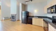 This two-story unit is defined by a modern, open design with light wood flooring throughout the main living areas and abundant natural light. The living room is a standout space, anchored by floor-to-ceiling wood-framed windows that flood the interior with sunlight and provide views of the surrounding urban streetscape. A balcony door opens directly from this area, extending the living space outdoors. A gas fireplace with a tiled surround adds warmth and character to the main level.

The kitchen is efficiently laid out with light maple cabinetry, dark granite-style countertops, and a full suite of black appliances including a refrigerator, dishwasher, electric range, and built-in microwave. Track lighting overhead keeps the kitchen well-illuminated. A dedicated utility area houses a stacked washer and dryer for in-unit convenience.

The bathroom features a light wood vanity with integrated sink, a large mirror, and dark tile flooring that extends into the hallway. Brushed nickel hardware and a towel ring add clean, contemporary detail.

Bedrooms are carpeted for comfort and each offer sliding wood-panel closet doors with built-in shelving and hanging rods. Windows allow natural light into these rooms, contributing to a bright and airy atmosphere throughout the unit.