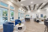 Located at 1521 Lee Rd in Douglasville, Georgia, this single-floor community clubhouse at Novo Sweetwater offers a well-appointed and versatile gathering space designed for both relaxation and socializing. The interior is finished with warm wood-look flooring throughout, crisp white walls, and soaring ceilings that lend an open, airy feel to the entire facility.

The main lounge and recreation area is a standout, featuring exposed dark wood ceiling beams, recessed lighting, and a full-size pool table positioned at the center of the room. Comfortable seating arrangements with plush blue accent chairs and a round coffee table create inviting conversation nooks. Floor-to-ceiling windows and glass-paneled doors flood the space with natural light and provide views of the outdoor pool area.

The entry zone is bright and welcoming, complete with a branded entry mat, built-in cabinetry with a beverage station, wall-mounted television, and high-top bar seating. A banquette-style dining area with marble-top tables lines one wall, complemented by a curated gallery of colorful abstract artwork.

The kitchenette area is thoughtfully designed with gray shaker cabinetry, a blue glass tile backsplash, open shelving, a stainless steel refrigerator, and a live-edge wood communal dining table — ideal for casual gatherings or resident events.
