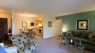 This single-floor residence offers a thoughtfully arranged open-concept layout that seamlessly connects the living room, dining area, and kitchen into one cohesive living space. The living room is carpeted and features a soothing color palette, highlighted by a bold green accent wall that adds a touch of personality and visual depth to the room. A glass-top coffee table and cushioned seating create a comfortable and welcoming atmosphere. Adjacent to the living area, the dining space accommodates a full-sized dining table and chairs, illuminated by a classic pendant light fixture that anchors the area with warmth. A framed chalkboard and wall art add decorative character to the dining nook. The kitchen is efficiently designed with oak-toned cabinetry, a gas range, and ample counter space, making it both practical and functional. The open sightlines between all three areas allow for easy interaction and a sense of spaciousness throughout. Neutral wall tones, consistent carpeting underfoot, and warm ambient lighting contribute to an overall cozy and livable ambiance, making this space well-suited for everyday comfort and casual entertaining.
