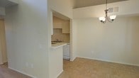 This single-floor space offers a practical and cohesive layout that connects the kitchen and dining area in an open, flowing arrangement. The kitchen is neatly tucked into a recessed alcove, defined by a pass-through counter that doubles as a breakfast bar. It features upper cabinetry in a light cream finish, a built-in sink, and a white dishwasher, providing functional storage and workspace within a compact footprint. The neutral beige walls and warm-toned tile flooring run consistently throughout, creating a unified, clean aesthetic. The dining area benefits from generous vertical space, with tall walls that lend an airy feel to the room. A classic chandelier with a bronze finish hangs above, adding a subtle decorative touch and providing ambient lighting. The open connection between the kitchen and dining zone enhances the sense of space, making the layout well-suited for everyday living. Overall, the space presents a simple, understated interior with a warm, neutral palette and practical design elements throughout.