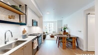 This well-appointed one-bedroom, one-bathroom residence showcases a modern design with a smart, efficient layout across a single floor. Light wood flooring flows throughout, creating a warm and cohesive aesthetic. The open-concept kitchen and living area serves as the heart of the home, featuring crisp white upper cabinetry with a geometric tile backsplash, two-tone lower cabinets in a warm gray, white quartz countertops, and stainless steel appliances including a gas range. A dining area with a round table sits adjacent to the kitchen, while the living space is anchored by a large window offering urban views and generous natural light. A built-in desk nook with floating shelves provides a dedicated workspace. The bedroom is bright and serene, with dual windows framing city and hillside views, continuing the light wood flooring. The bathroom features a single vanity with wood cabinetry, a large mirror, and matching flooring. A dedicated laundry area houses a full-size stacked washer and dryer with overhead cabinetry. The entry hallway offers additional storage, completing a residence that balances practicality with clean, contemporary style.