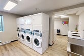 Located at 8717 Riverside Dr E in Windsor, Ontario, this shared laundry facility is a clean, efficiently designed space that provides residents with all the essentials for a convenient laundry experience. The room is finished with light wood-look flooring and neutral warm-grey walls, giving it a bright and tidy appearance throughout.

The main laundry area is anchored by a row of stacked front-loading washers and dryers — commercial-grade white units equipped with digital control panels — offering ample capacity for multiple users. Overhead, flush-mounted fluorescent lighting ensures the space is well-lit at all times. A stainless steel folding table runs along one wall, providing generous surface area for sorting and folding laundry.

An adjoining alcove serves as a utility and waiting area, featuring an additional stainless steel folding table, a chair, and a bulletin board mounted to the wall for resident notices. A stainless steel utility sink is also present near the entrance corridor, alongside a Sparkle payment kiosk for convenient card-based machine operation.

The entrance hallway is neatly appointed with informational signage, a fire safety station, and a potted plant that adds a welcoming touch. The space is designed with practicality and cleanliness in mind, making it a functional and comfortable shared amenity for building residents.