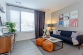 This thoughtfully designed single-floor residence offers a clean, contemporary aesthetic throughout, with crisp white walls, light wood-look flooring, and an efficient, well-organized layout. The bedroom is a comfortable retreat, featuring plush carpeting, tall ceilings, and a generous window that invites natural light. Framed artwork adds a personal touch, and a dedicated open wardrobe area provides practical storage. The kitchen is a standout feature, offering a galley-style configuration complemented by a central island with a dark countertop and pendant lighting above. White cabinetry lines one wall, paired with dark countertops and stainless steel appliances including a refrigerator, dishwasher, and range. Open shelving adds additional storage and display space. Light wood-look flooring flows seamlessly through the kitchen and entry area, where a wall-mounted coat rack provides a convenient drop zone. The bathroom is clean and functional, featuring a full soaking tub with a shower, neutral tile-look flooring, and bright white finishes throughout. A dedicated utility area houses a stacked washer and dryer alongside a water heater and HVAC unit, keeping all mechanical systems neatly contained. The overall ambiance is bright, modern, and move-in ready.