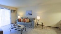 This single-floor residential space is located in the United States and offers a calm, well-organized living environment with a consistent neutral color palette throughout. The living area serves as the heart of the home, featuring soft wall-to-wall carpeting that adds warmth and comfort underfoot. The room is anchored by a generously sized slipcovered sofa accented with warm-toned throw pillows, complemented by glass and metal accent tables that lend a clean, contemporary feel to the space. Ambient lighting is provided by matching table lamps positioned on either side of the seating area, creating a warm and inviting atmosphere. A wall-mounted climate control unit ensures year-round comfort, while the smooth, light-colored walls offer a neutral backdrop that makes the space feel open and airy. Subtle decorative touches, including framed wall art, add a personal character to the room without overwhelming its understated aesthetic. The overall ambiance is practical yet comfortable, presenting a move-in-ready living space with a cohesive, relaxed design sensibility suited for everyday living.