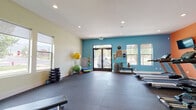 This single-floor fitness center is thoughtfully designed to support a full range of workout activities, divided into distinct functional zones that cater to both cardio and strength training needs. The space is characterized by vibrant color-blocked walls — a bold teal accent wall frames the main entrance area, while a striking orange feature wall with motivational typography reading "Energy, Lifestyle, Fitness, Strength, Work Out" energizes the cardio zone. Durable rubber flooring runs throughout, providing safety and comfort for all types of exercise. The cardio area is equipped with treadmills, an elliptical trainer, and a stationary bike, all positioned facing wall-mounted flat-screen televisions. The free weights and functional training zone offers a dumbbell rack, kettlebells, medicine balls, stability balls, foam rollers, and a weight bench. The strength training section features a multi-functional cable machine, additional benches, and a dumbbell rack, complemented by a mirrored wall that amplifies the sense of space. Recessed lighting illuminates the entire facility, while multiple large windows with roller shades invite natural light and outdoor views. Double glass entry doors complete the welcoming, energetic atmosphere of this well-appointed fitness facility.