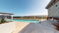This rooftop amenity space offers a well-appointed outdoor pool deck designed for both recreation and relaxation. The centerpiece is a generously sized lap pool with crystal-clear water, framed by large-format neutral-toned tile that extends across the entire deck surface, providing a clean, modern aesthetic. A black metal safety fence borders the perimeter, ensuring security while preserving sweeping panoramic views of the surrounding mountain range and urban landscape below — a standout feature that enhances the open-air atmosphere.

Access to the pool deck is provided through floor-to-ceiling glass doors with sleek dark metal framing, creating a seamless visual transition between the interior and exterior spaces. The entry area is well-lit with abundant natural light, and landscaped planters with ornamental grasses add a touch of greenery near the entrance. Informational and safety signage is clearly posted at the entry point, covering pool capacity, rules, and emergency procedures.

The deck itself offers ample open space around the pool for lounging, and the clean architectural lines of the surrounding structure — featuring white stucco walls and modern angular overhangs — complement the minimalist design language. The space benefits from open sky exposure, making it bright and airy throughout the day.