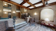 This distinguished lobby is a showcase of classic architectural craftsmanship, combining historic character with functional design. The space is defined by its exposed brick walls, which stretch upward to impressively high ceilings adorned with richly detailed coffered wood beams and paneling. Elegant brass chandeliers hang overhead, complementing the warm, ambient wall sconces that illuminate the space throughout.

The flooring features light-toned wood-look tile that contrasts beautifully with the warm brick tones, creating a refined yet welcoming atmosphere. A grand arched brick doorway fitted with ornate wrought-iron metalwork serves as a striking architectural focal point, while a secondary arched brick alcove adds further decorative depth to the walls.

Stained glass windows with colorful diamond-patterned panes allow natural light to filter warmly into a seating alcove, where wingback and upholstered armchairs invite visitors to rest. Additional seating with green upholstered chairs is positioned throughout the main area, catering to occupants and guests alike.

A bank of mailboxes and an upper-level mezzanine with decorative iron railings further highlight the building's multi-level character. An elevator provides convenient access between levels. The overall ambiance is one of timeless elegance, blending historic architectural detail with practical, well-appointed amenities.