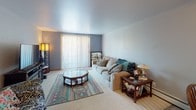 This single-floor space is thoughtfully organized into distinct living areas, each with its own character and function. The living room is a comfortable, carpeted space with soft, neutral-toned walls painted in a muted blue-gray. A large sliding glass door fitted with vertical blinds allows natural light to filter in, brightening the room. A cushioned sofa, glass-topped octagonal coffee table, and a patterned area rug contribute to its relaxed, welcoming atmosphere.

Adjacent to the living room, the dining area features a warm wood dining table with Windsor-style chairs and a bench, accented by decorative wall-mounted utensils and framed artwork. A ceiling fan with a light fixture overhead provides both ventilation and ambient lighting, while a freestanding shelving unit displays kitchen accessories and décor.

The kitchen is efficiently laid out with warm oak cabinetry, tile backsplash, and white appliances including a full-size refrigerator and dishwasher. Light vinyl flooring and a patterned accent rug complete the functional workspace.

The bedroom offers a calm retreat, featuring carpeted flooring, a full-size bed with colorful bedding, decorative wall art, and a window with drape curtains allowing soft natural light. The space is cozy and well-appointed with bedside furnishings.
