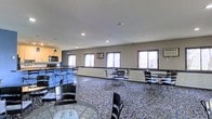 This single-floor common area is designed to accommodate a variety of gathering and dining needs. The open layout is furnished with multiple round and rectangular tables paired with black metal chairs, arranged to encourage both small group conversations and larger social gatherings. A textured, patterned carpet covers the floor throughout the main area, lending warmth and visual interest to the space.

Along one wall, a kitchenette is equipped with wood-toned cabinetry, a microwave, and countertop space, offering convenient access to basic food preparation. A series of large windows lines the perimeter, inviting generous amounts of natural light and providing views of the surrounding outdoor landscape. A wall-mounted air conditioning unit ensures climate comfort year-round.

The ceilings feature a classic textured finish with recessed lighting that supplements the natural light, maintaining a bright and welcoming ambiance throughout the day. The neutral gray walls provide a clean, understated backdrop that keeps the focus on the functionality and openness of the room. Overall, this community space is well-suited for hosting events, casual meetings, or everyday dining, offering a comfortable and practical environment for its users.