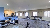 This single-floor common area is designed to accommodate a variety of gathering and dining needs. The open layout is furnished with multiple round and rectangular tables paired with black metal chairs, arranged to encourage both small group conversations and larger social gatherings. A textured, patterned carpet covers the floor throughout the main area, lending warmth and visual interest to the space.

Along one wall, a kitchenette is equipped with wood-toned cabinetry, a microwave, and countertop space, offering convenient access to basic food preparation. A series of large windows lines the perimeter, inviting generous amounts of natural light and providing views of the surrounding outdoor landscape. A wall-mounted air conditioning unit ensures climate comfort year-round.

The ceilings feature a classic textured finish with recessed lighting that supplements the natural light, maintaining a bright and welcoming ambiance throughout the day. The neutral gray walls provide a clean, understated backdrop that keeps the focus on the functionality and openness of the room. Overall, this community space is well-suited for hosting events, casual meetings, or everyday dining, offering a comfortable and practical environment for its users.