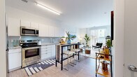 Located at 27 S King Street in Hampton, Virginia, this single-floor unit offers a thoughtfully designed open-concept layout that seamlessly connects the kitchen, dining, and living areas. The kitchen is outfitted with sleek white cabinetry, granite countertops, a light gray subway tile backsplash, and a full suite of stainless steel appliances, including a refrigerator, range, over-the-range microwave, and dishwasher. A bar-height table positioned at the kitchen's edge provides a versatile dining option that flows naturally into the living space.

The living area is bright and airy, benefiting from large sliding glass doors that invite natural light and offer views of the surrounding urban streetscape. Light-toned hardwood-style flooring runs throughout, contributing to the cohesive, contemporary aesthetic of the space.

The bedroom is a comfortable retreat featuring windows with horizontal blinds that frame exterior views of the city. A full bathroom includes a soaking tub with a shower, light tile flooring, and granite-topped vanity. A dedicated in-unit laundry area with a stacked washer and dryer adds everyday convenience. The overall design is clean, modern, and well-appointed throughout.