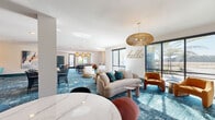 This contemporary single-floor amenity space is thoughtfully designed to foster both relaxation and productivity. The open-plan layout is anchored by a bold, patterned blue carpet that flows throughout, lending a cohesive and energetic aesthetic to the entire area. A welcoming lounge zone greets visitors with a curved cream sofa, colorful accent pillows, and warm cognac leather seating, all positioned beneath statement woven rattan pendant lights. A playful "hello" wall script adds a welcoming, personable touch, while large windows frame views of lush outdoor greenery and flood the space with natural light.

A casual dining and bar-height seating area accommodates social gatherings, complemented by a marble-top table and mixed modern chairs. Nearby, a sleek credenza with a decorative herringbone tile accent wall serves as a refined focal point. A secondary lounge offers a tufted leather sectional, a round marble dining table, and direct access to an outdoor balcony. The dedicated conference room features a long white table, modern rolling chairs, a textured teal accent wall, and a large window overlooking palm trees. Private computer workstations with decorative laser-cut partitions and woven wall sconces complete the space, offering focused, semi-enclosed work areas ideal for individual productivity.