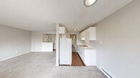 Located in Tacoma, WA, this one-bedroom, one-bathroom unit offers a thoughtfully arranged single-floor layout with a neutral color palette and clean finishes throughout. The open living area is generously proportioned, featuring soft carpeting, a textured ceiling, and built-in open shelving that adds functional character to the space. The galley-style kitchen runs alongside the living area and is equipped with white cabinetry, a white refrigerator, a single-basin sink, and wood-look flooring that contrasts neatly with the carpeted living zones. A hallway connects the main living space to the private areas of the unit, leading to the bedroom and bathroom. The bedroom is a comfortable size, carpeted, and features a window with blinds that allows natural light to filter in, along with baseboard heating and dual louvered bi-fold closet doors offering plentiful storage. The bathroom is well-appointed with a white vanity, white countertop, large mirror, and updated brushed hardware, with wood-look flooring continuing the cohesive design. A balcony provides an outdoor extension of the living space. The unit's consistent light gray tones and white trim create a bright, clean ambiance throughout.