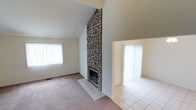 This two-story home makes an immediate impression with its open, vaulted living area, where soaring ceilings draw the eye upward and create a sense of generous vertical space. The standout architectural feature is a commanding floor-to-ceiling stone fireplace, constructed from natural stacked stone that anchors the main living area with both visual weight and rustic character. The hearth sits on a tiled surround that transitions into the carpeted living room floor, offering a clear delineation between comfort and function. Large windows fitted with vertical blinds allow natural light to filter into the space, brightening the neutral-toned walls and light-colored carpet. Adjacent to the main living area is an open, tiled space — likely a dining or entry area — also benefiting from natural light through its own window. The transition between carpet and tile flooring throughout the main level adds textural contrast and helps define the distinct functional zones. The high vaulted ceiling in the living room, paired with the dramatic stone chimney that rises to the peak, gives the space an airy, expansive quality. Overall, the home presents a classic, well-structured layout with timeless architectural details suited to comfortable everyday living.