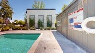 Located at 4729 Manzanita Ave in Carmichael, California, this outdoor amenity space centers around a generously sized, crystal-clear swimming pool that serves as the focal point of the area. The pool is framed by a wide concrete surround that provides ample space for lounging and movement, with multiple chaise lounge chairs and patio seating arranged throughout the deck. Shade umbrellas and a covered pergola structure with seating offer comfortable retreats from the California sun, making the space functional across all hours of the day. Vibrant green accent chairs add a pop of color against the neutral hardscape tones. The area is enclosed by a secure metal fence, ensuring safety and privacy, while mature trees surrounding the perimeter create a natural, shaded backdrop. Manicured landscaping with planters and shrubs lines the boundaries of the space, enhancing its visual appeal. A dedicated utility and facilities building is positioned on-site, complete with posted pool rules and safety signage. The multi-story residential buildings visible in the background provide context for the community setting. Under clear blue skies, this outdoor space delivers a resort-like atmosphere well-suited for relaxation and recreation.