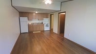 This single-floor space offers an open-concept layout that combines the kitchen and living area into one cohesive, functional space. The warm hardwood-style flooring runs throughout, lending a consistent and inviting aesthetic to the entire area.

The kitchen is equipped with medium-toned wood cabinetry offering generous storage, a double sink, granite-style countertops, a white electric range, and a full-size white refrigerator. An under-cabinet range hood is positioned above the cooktop, and a small utility mat rests near the sink area. Neutral-toned walls complement the cabinetry and flooring throughout.

The adjoining living area benefits from the open layout, creating a sense of spaciousness. A ceiling fan with a light fixture is centrally mounted, providing both airflow and ambient lighting. Crown molding-style trim accents the upper walls, adding a touch of architectural detail.

A hallway visible from the main area leads to additional rooms, suggesting a practical and straightforward floor plan. The overall ambiance is clean, functional, and well-maintained, suited for everyday living.