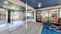 Located at 6000 Boca Raton Blvd in Fort Worth, TX, this fitness center offers a well-equipped workout space designed to support a comprehensive exercise routine. The room features a bold navy-painted ceiling accented by recessed lighting, creating a modern, energetic atmosphere that contrasts with the neutral gray walls. The flooring is a combination of striped gray carpet and vibrant blue area carpet, visually dividing the cardio and strength training zones.

The cardio section is furnished with treadmills, an elliptical trainer, and a stationary recumbent bike, all positioned near large windows that invite natural light and provide views of the surrounding outdoor area. The strength training zone includes a cable machine, an adjustable weight bench, a dumbbell rack with multiple weights, a rowing machine, and a stability ball, offering a diverse range of workout options. Floor-to-ceiling mirrors line one wall, enhancing the sense of openness and allowing users to monitor their form during exercise.

A glass-panel door connects the fitness center to an adjacent common area, while a separate exterior door and multiple windows ensure the space feels bright and accessible. The layout is thoughtfully organized, making efficient use of the available floor area to accommodate a full-body fitness experience.