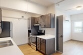 This 2-bedroom, 1-bathroom unit offers a practical and modern layout across a single floor. Light wood-look flooring runs throughout the entire space, providing a clean and cohesive aesthetic. The galley-style kitchen is a standout feature, equipped with dark-toned cabinetry, white countertops, stainless steel appliances including an electric range and refrigerator, a double basin sink, and a built-in microwave. Ample upper and lower cabinet storage lines both sides of the kitchen, maximizing functionality in the compact footprint. Each bedroom is bright and simply finished with white walls, blinds-covered windows that offer views of surrounding greenery, and wall-mounted mini-split HVAC units for individualized climate control. The primary bedroom includes a double-door closet and a ceiling fan, while the second bedroom offers a comparably airy feel with natural light. The bathroom features light wood-look flooring, a walk-in shower with a curved curtain rod, a white vanity with countertop, a medicine cabinet, and matte black hardware and fixtures. Exposed conduit and industrial-style ceiling details add a subtle urban character to the overall design of the unit.