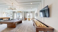 Located in Orlando, FL, this well-appointed community game room offers a thoughtfully designed recreational space that blends contemporary style with comfortable functionality. The room is anchored by a full-size pool table and a wooden shuffleboard table, providing a variety of entertainment options for residents and guests alike. A curved, channel-tufted banquette in teal and cognac upholstery lines one wall, paired with round pedestal tables to create a relaxed lounge area ideal for socializing. The coffered ceiling detail, adorned with decorative molding, adds architectural dimension overhead, while a striking brass Sputnik-style chandelier serves as the room's centerpiece, casting warm ambient light throughout. Gold-toned wall sconces complement the chandelier and highlight a curated gallery wall of framed artwork. Large windows dressed with sheer patterned curtains invite abundant natural light and offer views of the surrounding landscape. Wall-mounted flat-screen televisions are positioned for easy viewing from multiple vantage points. A built-in cabinetry unit with a wet bar area is also present, adding convenience to the entertaining space. The neutral-toned, geometric-patterned carpet grounds the room and ties together its modern coastal aesthetic, making this game room a vibrant and inviting hub for recreation and community engagement.