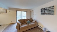 This single-floor, one-bedroom, one-bathroom unit offers a straightforward and functional layout designed to maximize usability within a compact footprint. The living room serves as the central gathering space, featuring wall-to-wall carpet, a large sliding glass door that fills the room with natural light and provides access to a balcony with outdoor views. A baseboard heating unit runs along the wall, and the entry door is conveniently located within this space.

Adjacent to the living area is a dedicated dining nook with tile-style flooring, a wall-mounted air conditioning unit, and overhead pendant lighting, creating a defined space for meals. The kitchen is efficiently arranged with wood-toned cabinetry, laminate countertops, a gas range, refrigerator, and a single-basin sink, offering all essential cooking amenities.

The bedroom is a comfortably sized, carpeted room with a textured ceiling, a window with curtains, and baseboard heating. It provides adequate space for standard bedroom furnishings. The bathroom includes a vanity with a wall mirror, overhead lighting, and a shower, finished with a textured wall treatment for added character.