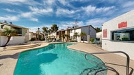 Located in Phoenix, Arizona, this outdoor community space centers around a generously sized freeform swimming pool, designed to evoke a resort-style atmosphere under the expansive Arizona sky. The pool's curvilinear shape creates a dynamic and inviting focal point, with crystal-clear turquoise water reflecting the abundant desert sunshine.

Surrounding the pool is a spacious concrete sun deck furnished with lounge chairs, patio tables, and shade umbrellas, offering ample room for relaxation and outdoor recreation. The deck is thoughtfully arranged to accommodate both sunbathers and social gatherings. A raised spa or wading area is situated adjacent to the main pool, adding an additional layer of amenity to the space.

Lush landscaping enhances the environment throughout, with mature palm trees and ornamental shrubs providing natural shade and a tropical aesthetic that complements the Phoenix climate. Decorative lamp posts are positioned around the perimeter, ensuring the area is well-lit during evening hours.

The pool area is enclosed by a wrought iron fence, providing both security and a sense of defined community space. Multi-story residential buildings frame the backdrop, their stucco exteriors and balconies contributing to the classic Southwestern architectural character. The overall ambiance is relaxed and functional, well-suited to the warm desert climate of the Phoenix metropolitan area.