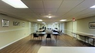 This single-floor multipurpose room offers a spacious, open layout well-suited for a variety of gatherings, meetings, or community events. The room features a drop-panel ceiling fitted with recessed fluorescent lighting that evenly illuminates the entire space, creating a bright and functional environment. The flooring consists of warm-toned hardwood-style planks that contrast with the neutral, light green-toned walls, which are accented by white chair rail molding running along the perimeter. Several framed artworks are hung at regular intervals, adding a modest decorative touch to the otherwise practical setting. The room is furnished with multiple long folding tables arranged in rows, accompanied by stackable chairs that can be easily reconfigured to accommodate different event formats. Additional stacked chairs are visible along the walls, indicating flexible seating capacity. An emergency exit sign is visible near the far end of the room, and a utility table is positioned along one side wall. The overall ambiance is clean, organized, and functional, making this space well-equipped to serve as a community hall, banquet room, classroom, or event venue.