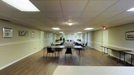 This single-floor multipurpose room offers a spacious, open layout well-suited for a variety of gatherings, meetings, or community events. The room features a drop-panel ceiling fitted with recessed fluorescent lighting that evenly illuminates the entire space, creating a bright and functional environment. The flooring consists of warm-toned hardwood-style planks that contrast with the neutral, light green-toned walls, which are accented by white chair rail molding running along the perimeter. Several framed artworks are hung at regular intervals, adding a modest decorative touch to the otherwise practical setting. The room is furnished with multiple long folding tables arranged in rows, accompanied by stackable chairs that can be easily reconfigured to accommodate different event formats. Additional stacked chairs are visible along the walls, indicating flexible seating capacity. An emergency exit sign is visible near the far end of the room, and a utility table is positioned along one side wall. The overall ambiance is clean, organized, and functional, making this space well-equipped to serve as a community hall, banquet room, classroom, or event venue.