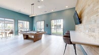 This contemporary community game room is thoughtfully designed for recreation and social interaction. The space is defined by bold teal-painted walls that contrast elegantly with a warm, natural wood plank accent wall, creating a vibrant yet inviting atmosphere. Large-format light tile flooring extends throughout, contributing to a clean, open feel.

At the center of the room sits a full-size wood-framed pool table, positioned beneath a sleek linear pendant light fixture that provides focused illumination. A wall-mounted flat-screen television is prominently displayed on the wood accent wall, accompanied by a built-in counter and bar-height seating — ideal for watching games or casual gatherings.

Abundant natural light floods the space through multiple floor-to-ceiling windows and glass doors along two walls, offering direct views of the outdoor courtyard and surrounding community buildings. A marble-top bistro table with upholstered chairs provides an intimate seating nook near the windows, perfect for conversation or casual dining.

Abstract artwork in warm earth tones adorns the teal walls, adding artistic character to the room. Glass double doors connect the space to adjacent amenity areas, enhancing the sense of openness and flow. Overall, the room balances modern design with functional recreational amenities in a well-lit, stylish environment.