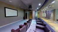 This space is designed as a private screening room, delivering a true cinema experience within a single-floor layout. The room is anchored by a large, wall-mounted projection screen at the front, paired with a ceiling-mounted projector positioned centrally overhead for optimal image quality. Rows of plush, dark leather recliner-style seats are arranged in a tiered formation, ensuring clear sightlines from every position. The seating is organized into two columns with a central aisle, and each seat features individual armrests and footrests for maximum comfort. The flooring is finished in a soft, neutral carpet that contributes to the room's acoustic qualities. Recessed lighting is integrated into the ceiling, providing adjustable ambient illumination. One side of the room features floor-to-ceiling glass partitioning, allowing visual connectivity to an adjacent space while maintaining acoustic separation. The walls are finished in muted, warm tones that complement the room's cinematic atmosphere. A vivid piece of artwork is visible through the glass partition, adding a decorative element. The overall design is polished and purposeful, combining comfort with high-quality audiovisual infrastructure.