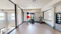 Located on N Oklahoma Ave in Oklahoma City, this single-floor fitness studio offers a generously sized, open layout designed to accommodate a wide range of exercise activities. The space is thoughtfully divided into two primary functional zones: an open movement area ideal for yoga, stretching, or group fitness classes, and a dedicated equipment zone stocked with medicine balls, kettlebells, stability balls, and additional free weights organized on structured racks.

The flooring throughout is a smooth, light-toned wood-look surface that provides both durability and aesthetic consistency. Large floor-to-ceiling windows with black metal framing line one side of the studio, flooding the interior with natural light and providing views of the surrounding outdoor balcony area. A set of glass-paneled doors offers direct access to the exterior.

Full-length wall mirrors are positioned in the equipment zone, enhancing the sense of space and allowing users to monitor their form during workouts. Recessed ceiling lighting supplements natural light, while a wall-mounted flat-screen television adds a multimedia element to the open movement area. The neutral color palette—white walls, teal baseboards, and light flooring—creates a clean, modern, and motivating environment throughout.