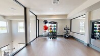Located on N Oklahoma Ave in Oklahoma City, this single-floor fitness studio offers a generously sized, open layout designed to accommodate a wide range of exercise activities. The space is thoughtfully divided into two primary functional zones: an open movement area ideal for yoga, stretching, or group fitness classes, and a dedicated equipment zone stocked with medicine balls, kettlebells, stability balls, and additional free weights organized on structured racks.

The flooring throughout is a smooth, light-toned wood-look surface that provides both durability and aesthetic consistency. Large floor-to-ceiling windows with black metal framing line one side of the studio, flooding the interior with natural light and providing views of the surrounding outdoor balcony area. A set of glass-paneled doors offers direct access to the exterior.

Full-length wall mirrors are positioned in the equipment zone, enhancing the sense of space and allowing users to monitor their form during workouts. Recessed ceiling lighting supplements natural light, while a wall-mounted flat-screen television adds a multimedia element to the open movement area. The neutral color palette—white walls, teal baseboards, and light flooring—creates a clean, modern, and motivating environment throughout.