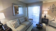 This single-floor residence offers a well-appointed living space designed for both comfort and functionality. The living room serves as the heart of the home, centered around a plush, tufted sofa adorned with decorative accent pillows in neutral tones. A large, framed wall mirror adds depth and dimension to the room, enhancing the sense of space. The textured walls provide a subtle visual interest that complements the overall aesthetic. Natural light floods the interior through a full-length sliding glass door fitted with vertical blinds, which opens directly onto a private outdoor balcony — ideal for relaxing or enjoying the surrounding environment. The balcony, visible through the glass door, features outdoor seating and is enclosed by a railing. A dark coffee table grounds the seating area, while a decorative centerpiece adds a finishing touch. The flooring transitions from carpet in the main seating area, contributing to a warm and cozy atmosphere throughout. The space features a textured ceiling and neutral wall tones that create a calm, cohesive ambiance, making it a versatile and inviting environment.